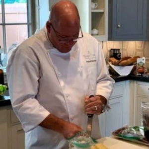 A man standing in a kitchen. He is looking down at the table in front of him where he is preparing a dish. He is wearing glasses and a white chef's jacket and is holding a grater in one hand. To his left and right there are bags filled with cooking ingredients.
