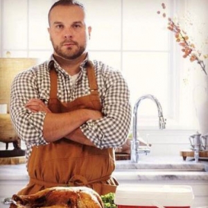 A man standing in a kitchen. He is wearing a light brown apron and his arms are folded at his chest. On the table in front of him is a cooked turkey.